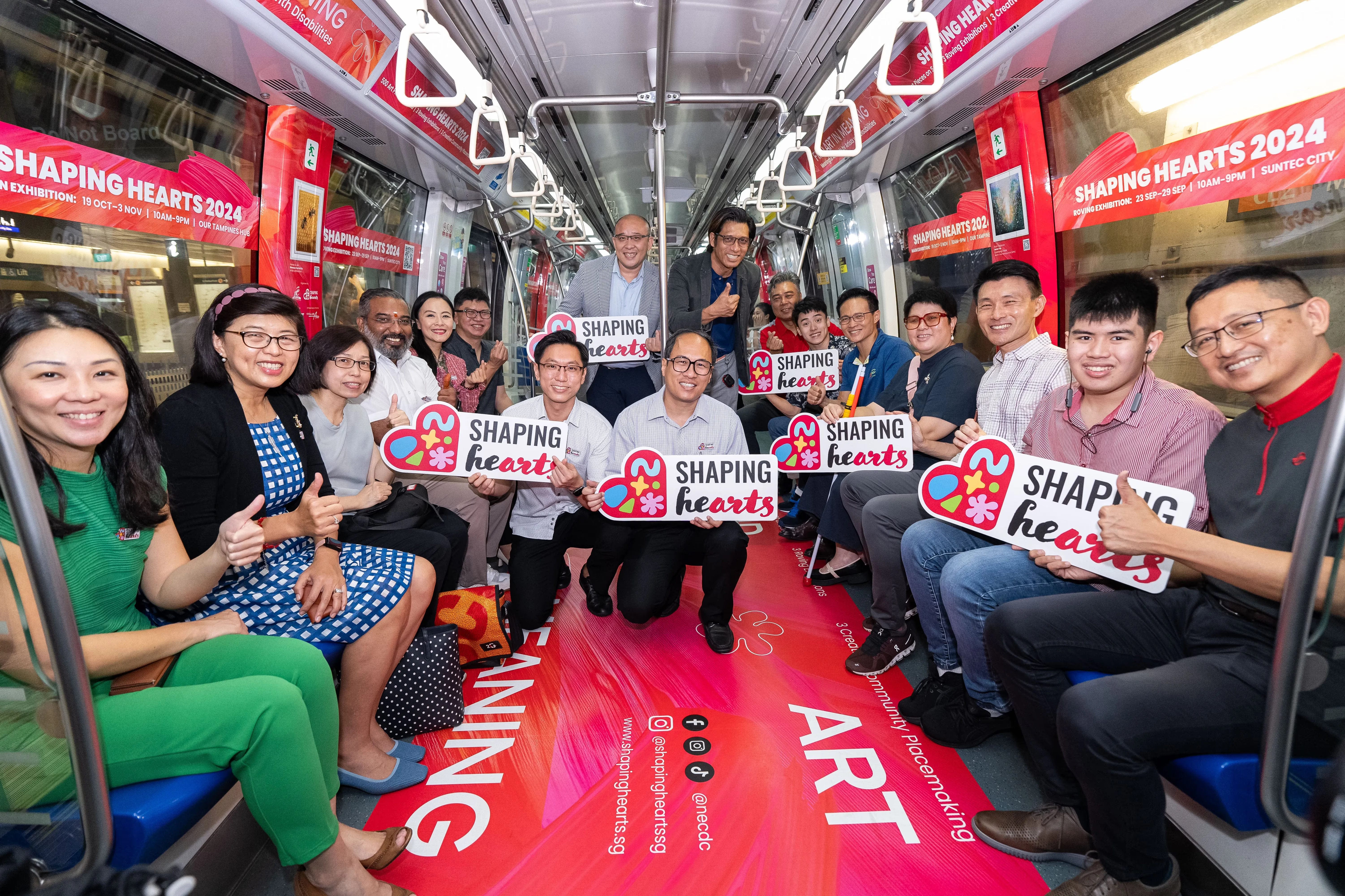 People on a train holding "Shaping Hearts" signs with red advertisements for "Shaping Hearts 2024" in the background.